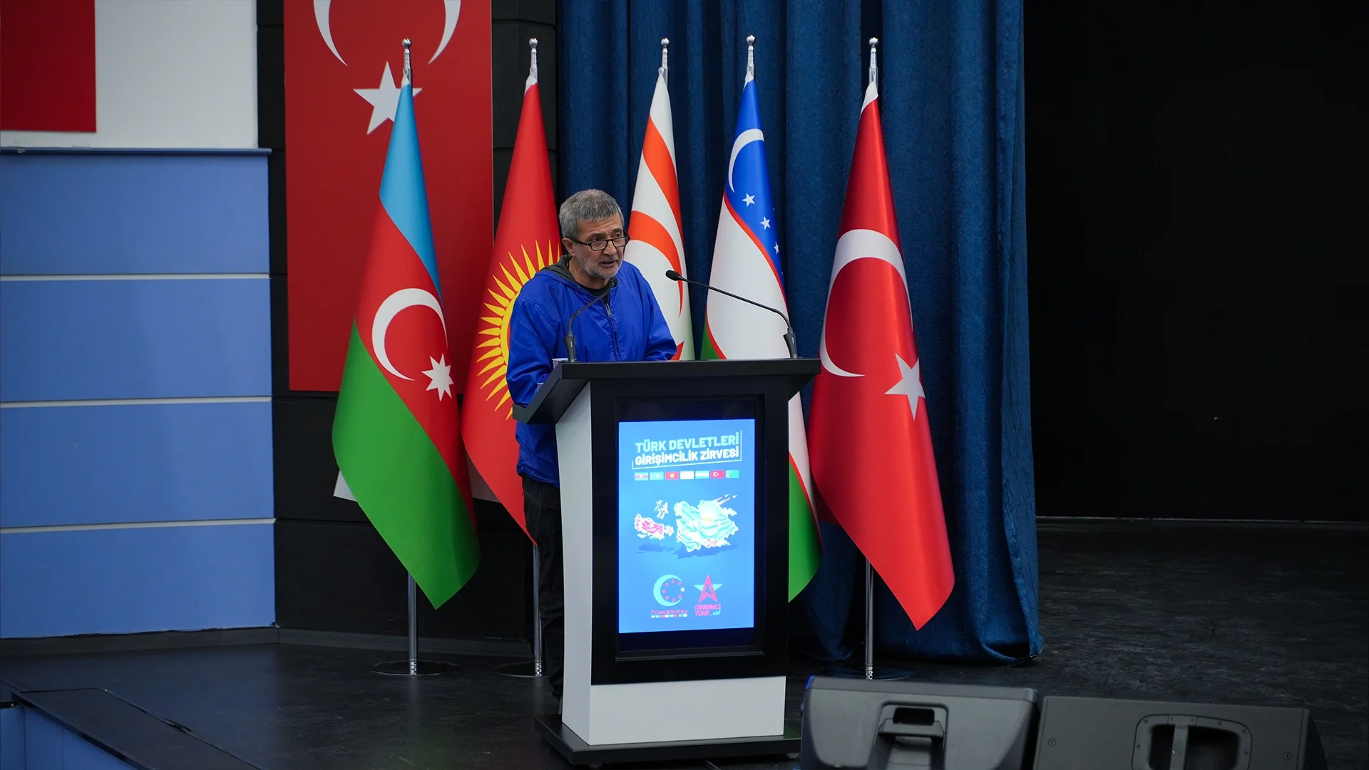Türk devletlerinin bayrakları önünde, “Türk Devletleri Girişimcilik Zirvesi” kürsüsünde konuşan gözlüklü bir adam