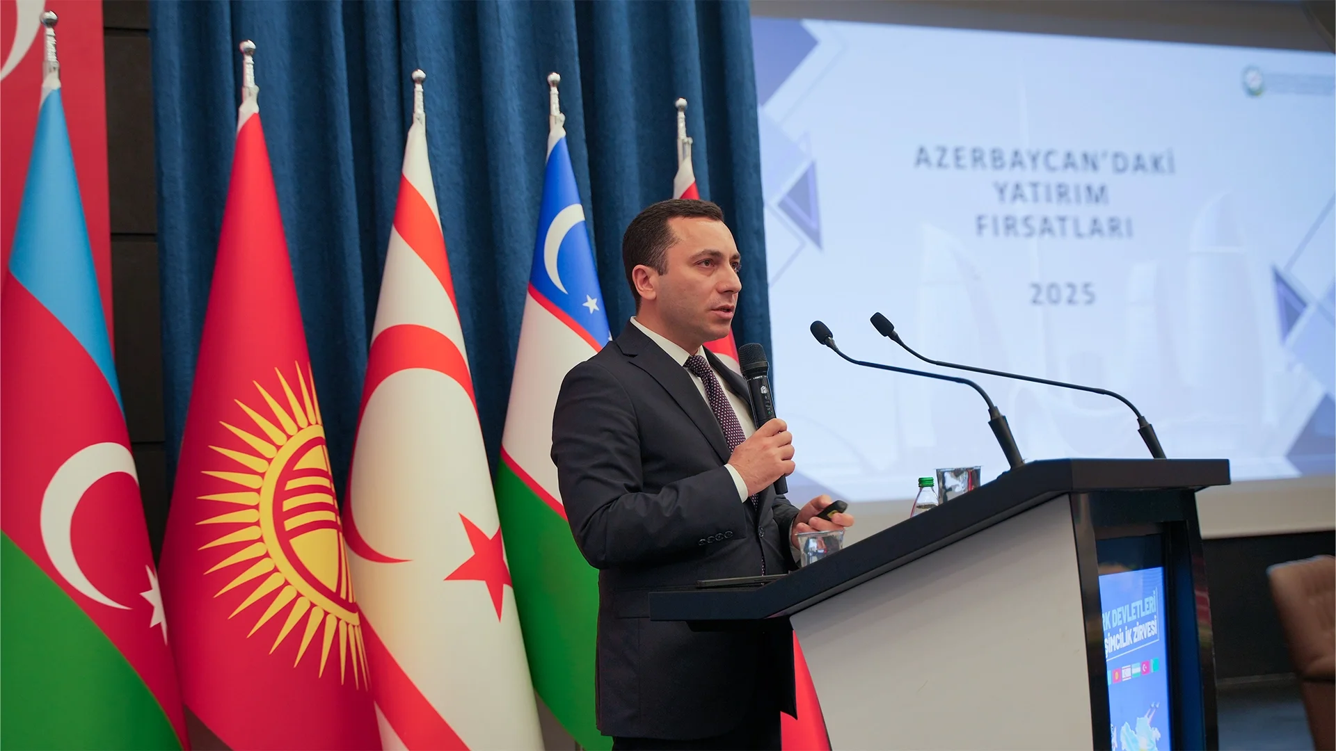 Türk devletlerinin bayrakları önünde kürsüde konuşan takım elbiseli bir adam; ekranda “Azerbaycan’daki Yatırım Fırsatları 2025” yazısı