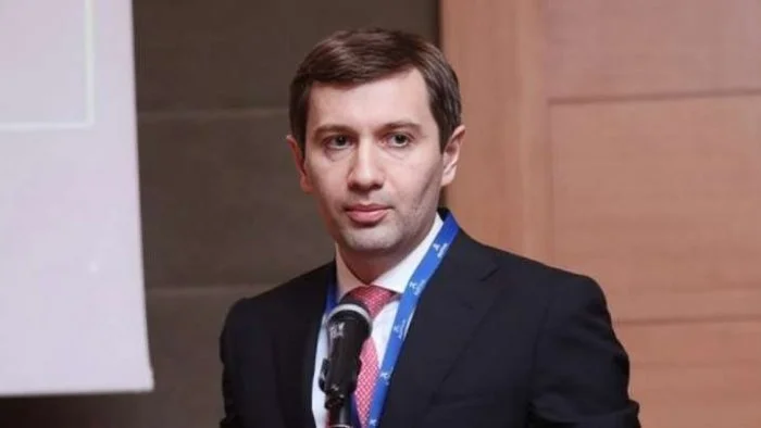 Yaka kartı takmış, takım elbiseli bir adam mikrofon önünde konuşuyor
