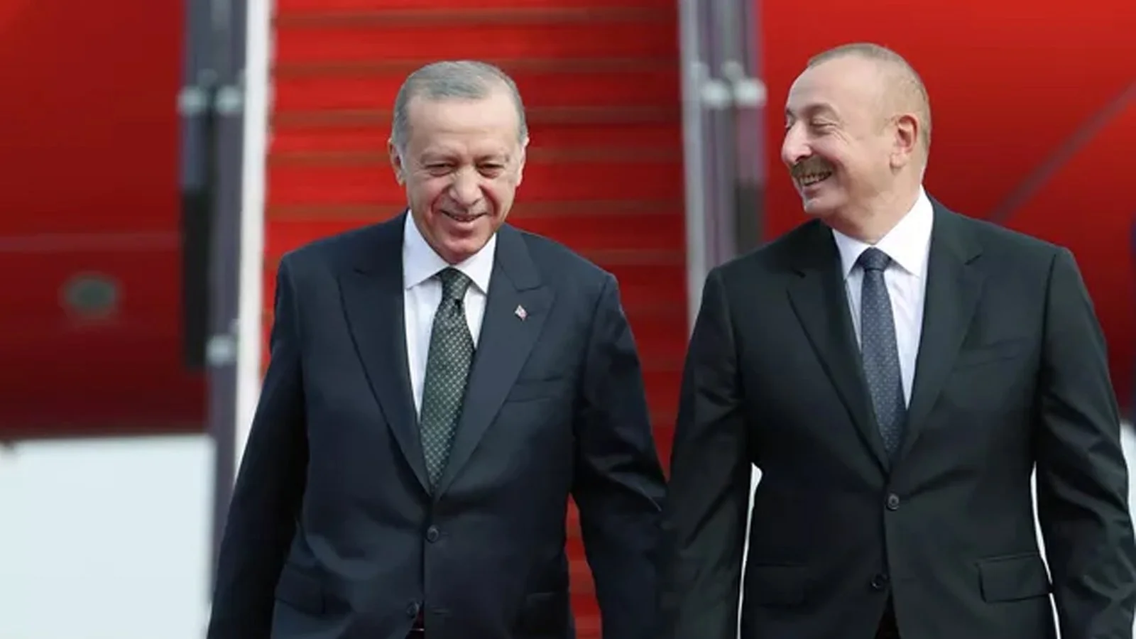 Kırmızı halı ve uçak merdiveni önünde gülümseyerek yan yana yürüyen takım elbiseli iki adam