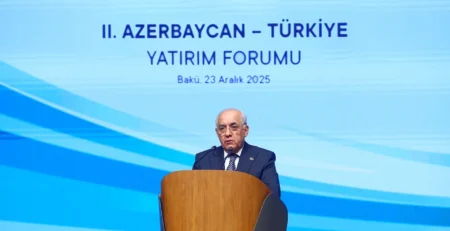 Mavi fon önünde kürsüde konuşan takım elbiseli bir adam; ekranda “II. Azerbaycan – Türkiye Yatırım Forumu, Bakü, 23 Aralık 2025” yazısı
