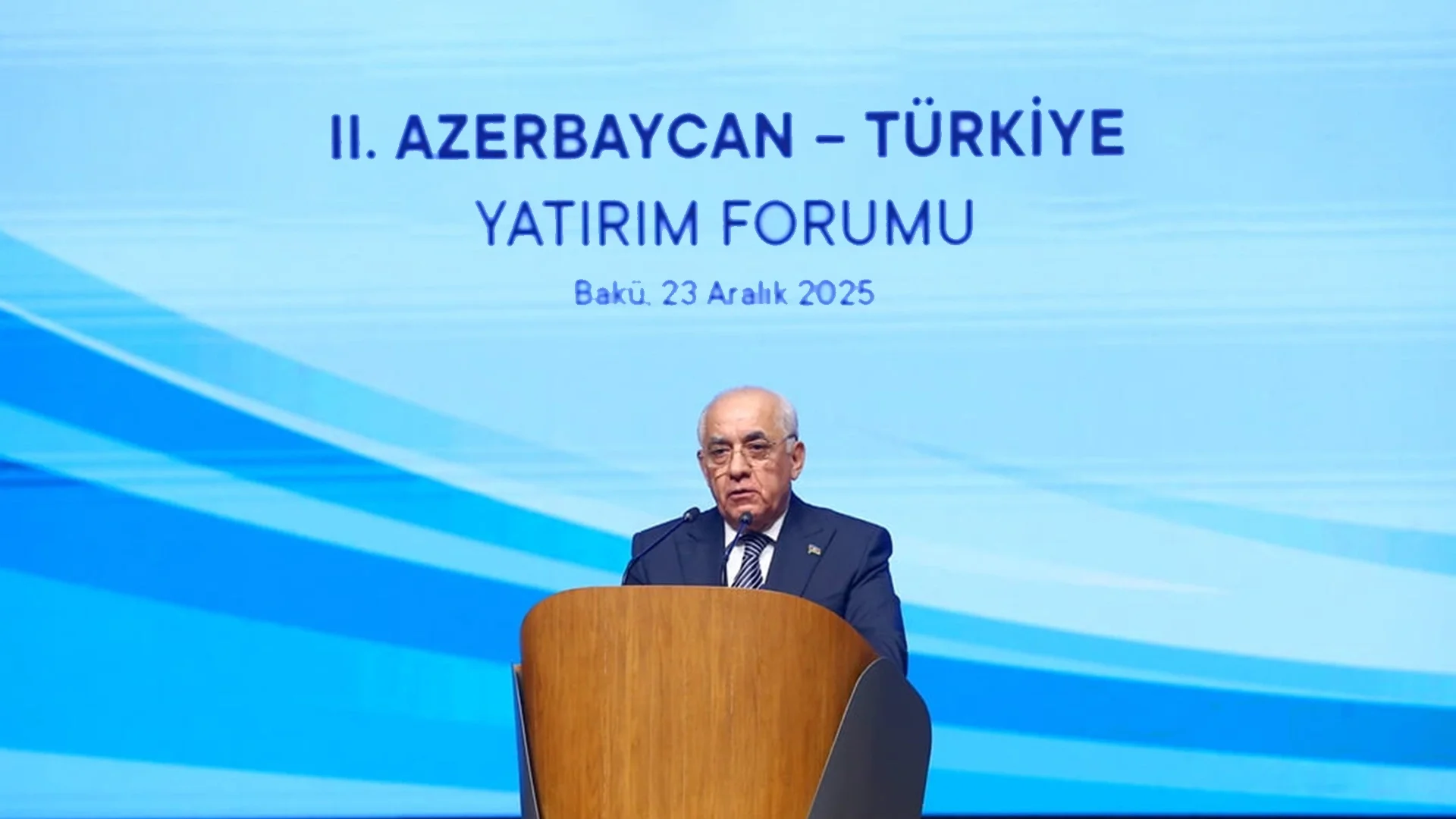 Mavi fon önünde kürsüde konuşan takım elbiseli bir adam; ekranda “II. Azerbaycan – Türkiye Yatırım Forumu, Bakü, 23 Aralık 2025” yazısı