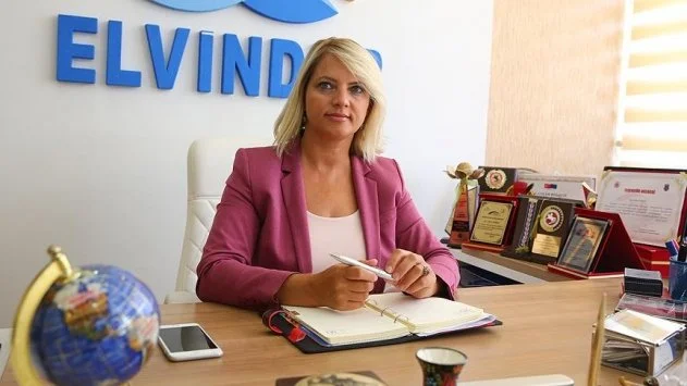 ELVİNDER logosu önünde, ödüllerle dolu ofisinde masa başında oturan pembe ceketli bir kadın