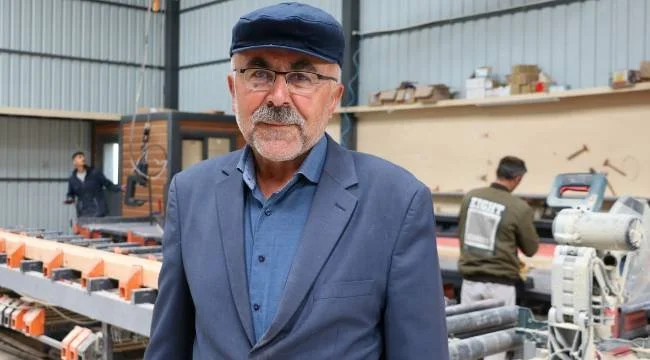 Kasketli, gözlüklü ve bıyıklı yaşlı bir adam; arkasında ahşap ev üretim atölyesinde çalışan işçiler