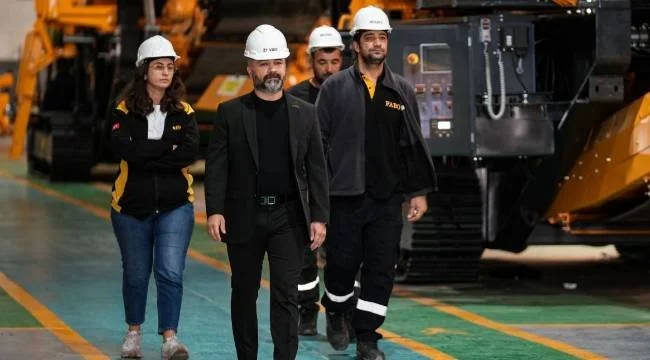 Fabrikada iş makineleri arasında yürüyen, baretli dört çalışan