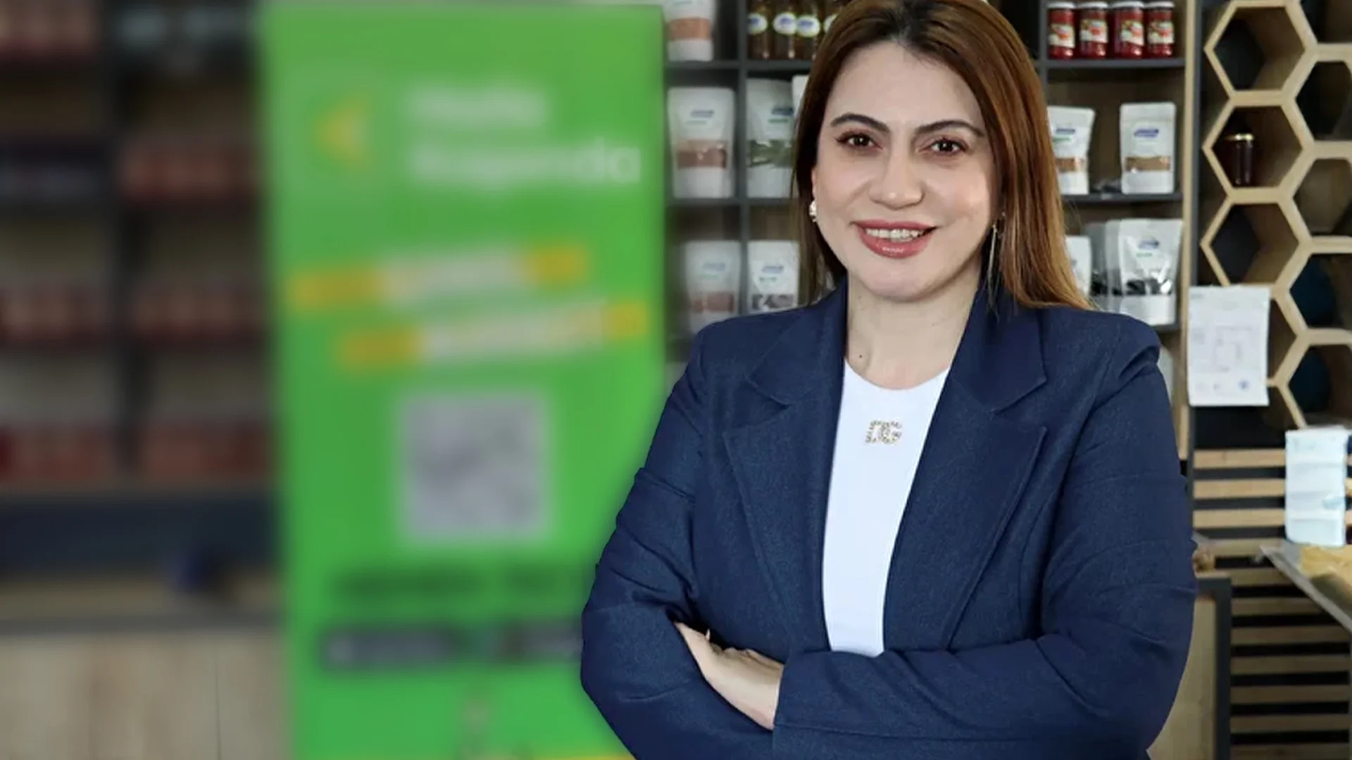 Kollarını kavuşturmuş, lacivert ceketli, gülümseyen bir kadın; arka planda raflarda yöresel ürünler ve yeşil tanıtım panosu