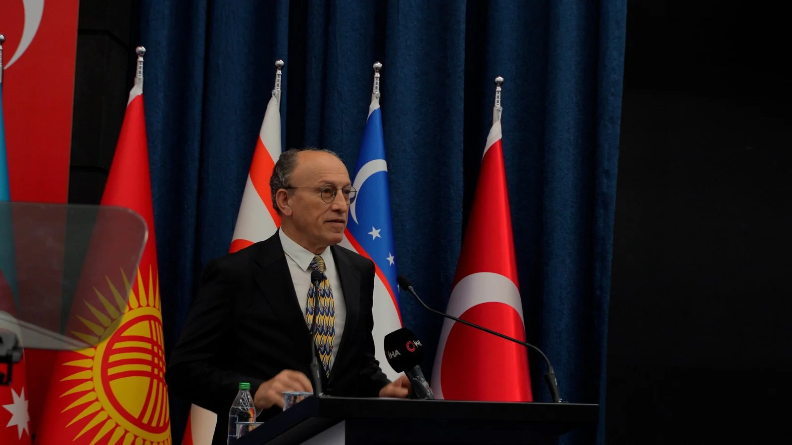 Kırgızistan, Türkiye ve Özbekistan bayrakları önünde kürsüde konuşan, gözlüklü, takım elbiseli bir adam