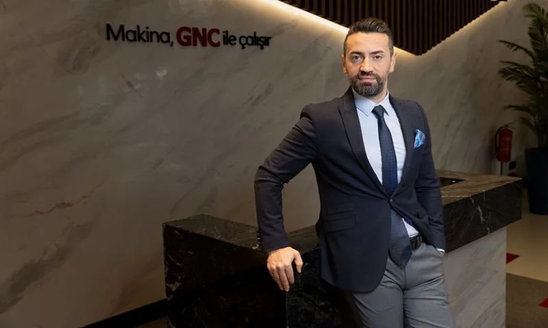 “Makina, GNC ile çalışır” yazılı mermer duvar önünde, resepsiyon tezgâhına yaslanmış lacivert takım elbiseli bir adam