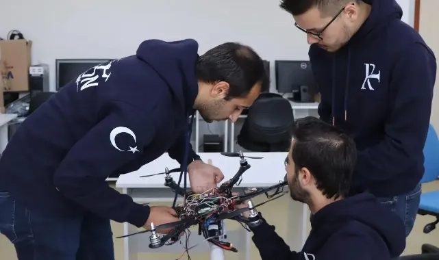 Laboratuvarda masa üzerindeki drone’u inceleyen, lacivert sweatshirt giymiş üç genç