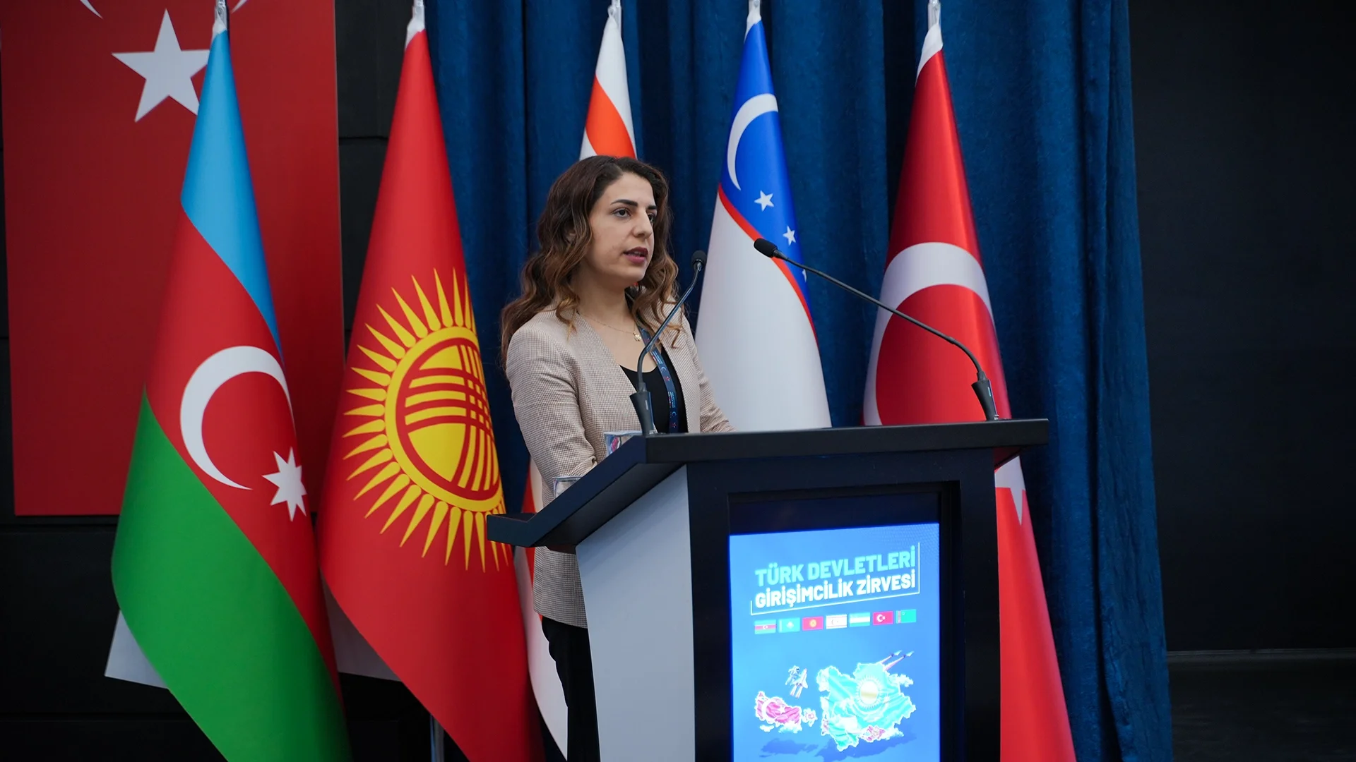 Türkiye, Azerbaycan, Kırgızistan ve Özbekistan bayrakları önünde, “Türk Devletleri Girişimcilik Zirvesi” kürsüsünde konuşan bir kadın