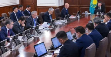 Kazakistan bayrağı bulunan toplantı salonunda, uzun masa etrafında tabletlerle oturan takım elbiseli yetkililer