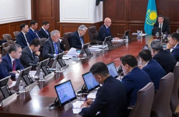 Kazakistan bayrağı bulunan toplantı salonunda, uzun masa etrafında tabletlerle oturan takım elbiseli yetkililer