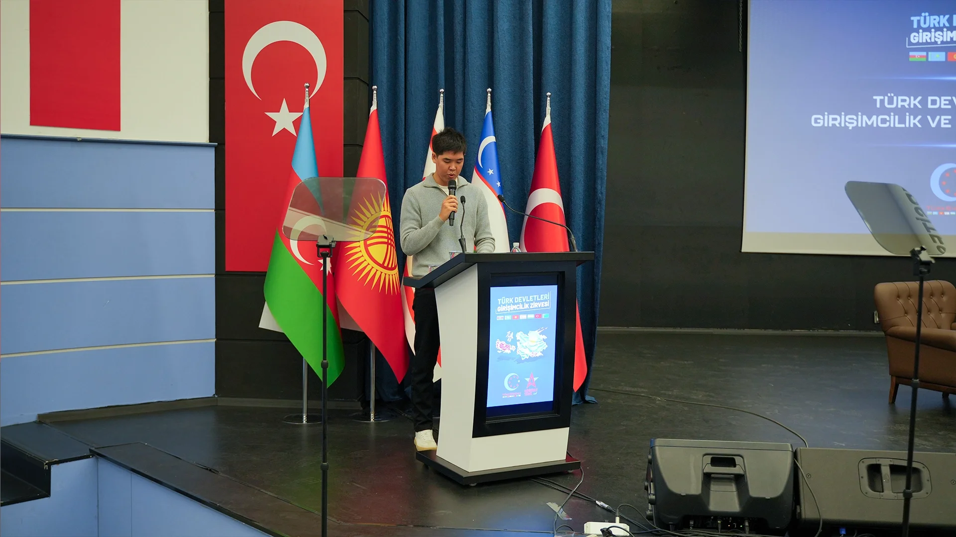 Türkiye, Azerbaycan, Kırgızistan ve Özbekistan bayrakları önünde, zirve kürsüsünde mikrofonla konuşan genç bir adam