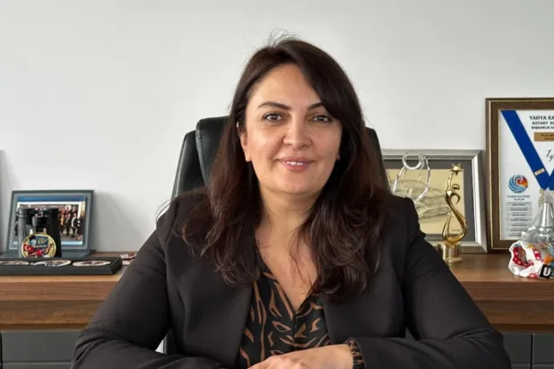Ofisinde masa başında oturan, koyu renk ceketli, gülümseyen bir kadın; arkada çerçeveli belgeler ve ödüller