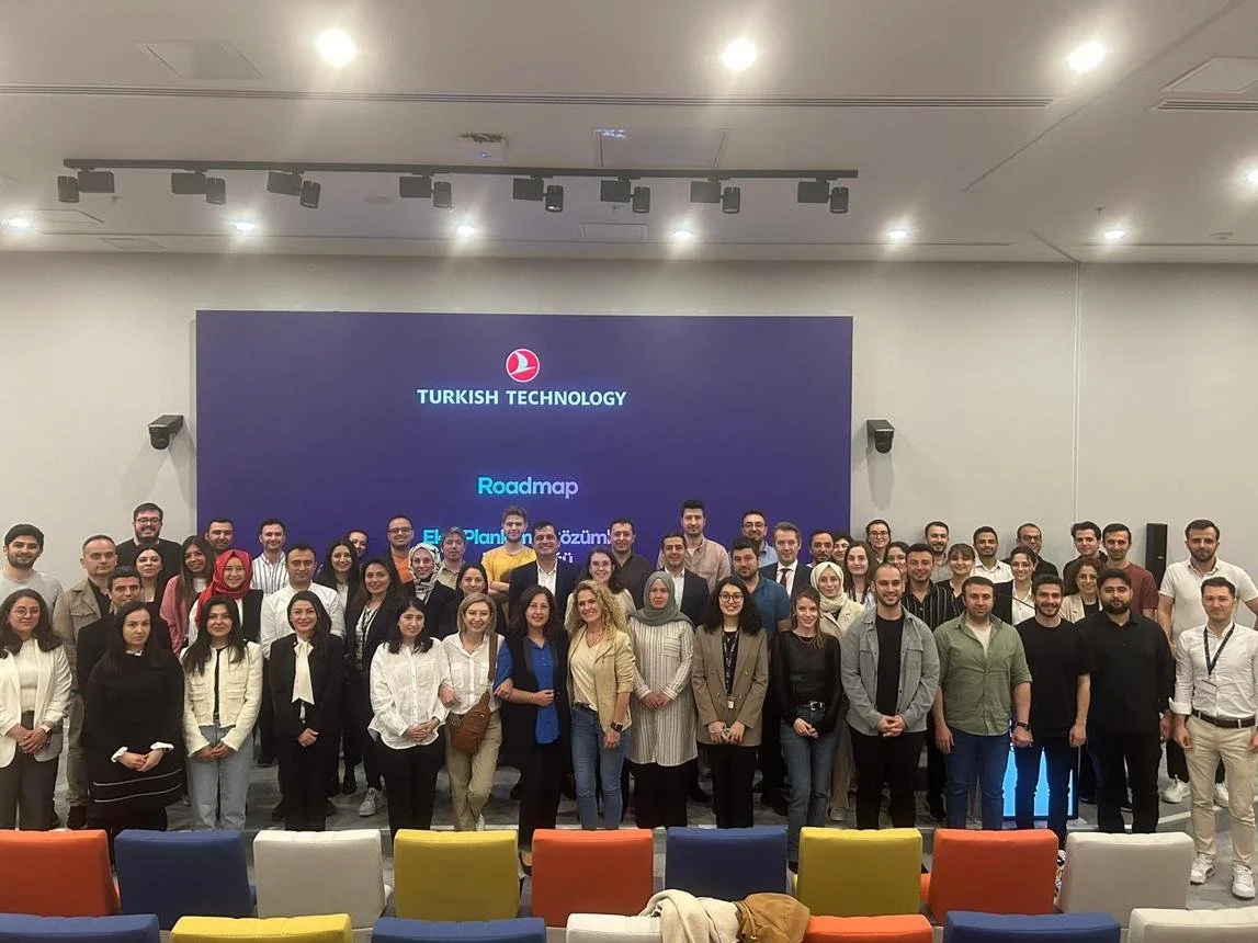Turkish Technology logolu ekranın önünde, renkli koltukların arkasında poz veren kalabalık bir katılımcı grubu