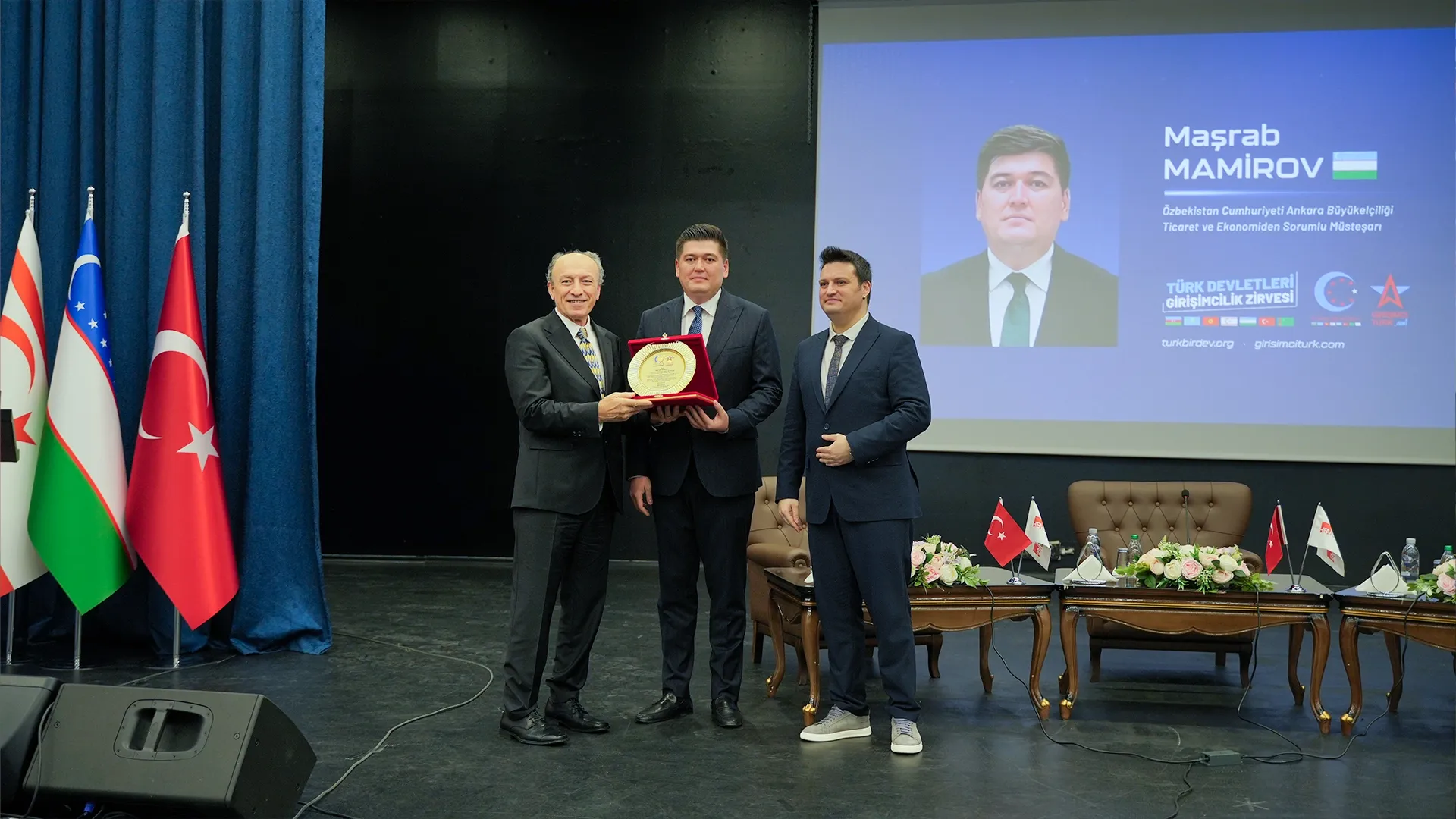 Türk devletlerinin bayrakları önünde sahnede plaket takdim eden üç adam; arkadaki ekranda “Maşrab Mamirov” yazısı