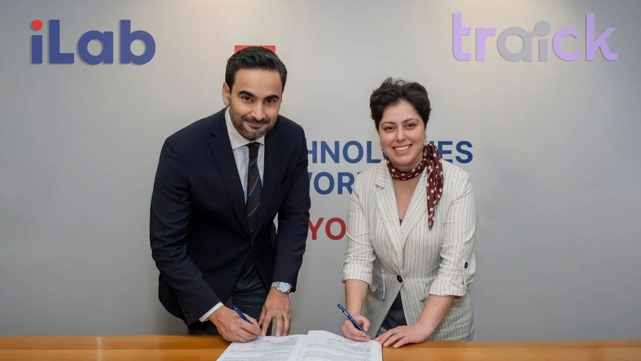 iLab ve Traick logolu fon önünde masada sözleşme imzalayan takım elbiseli bir adam ve bir kadın