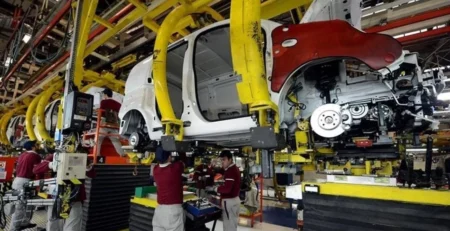 Otomobil fabrikasında sarı askı sistemlerine bağlı araç gövdeleri üzerinde çalışan bordo tulumlu işçiler