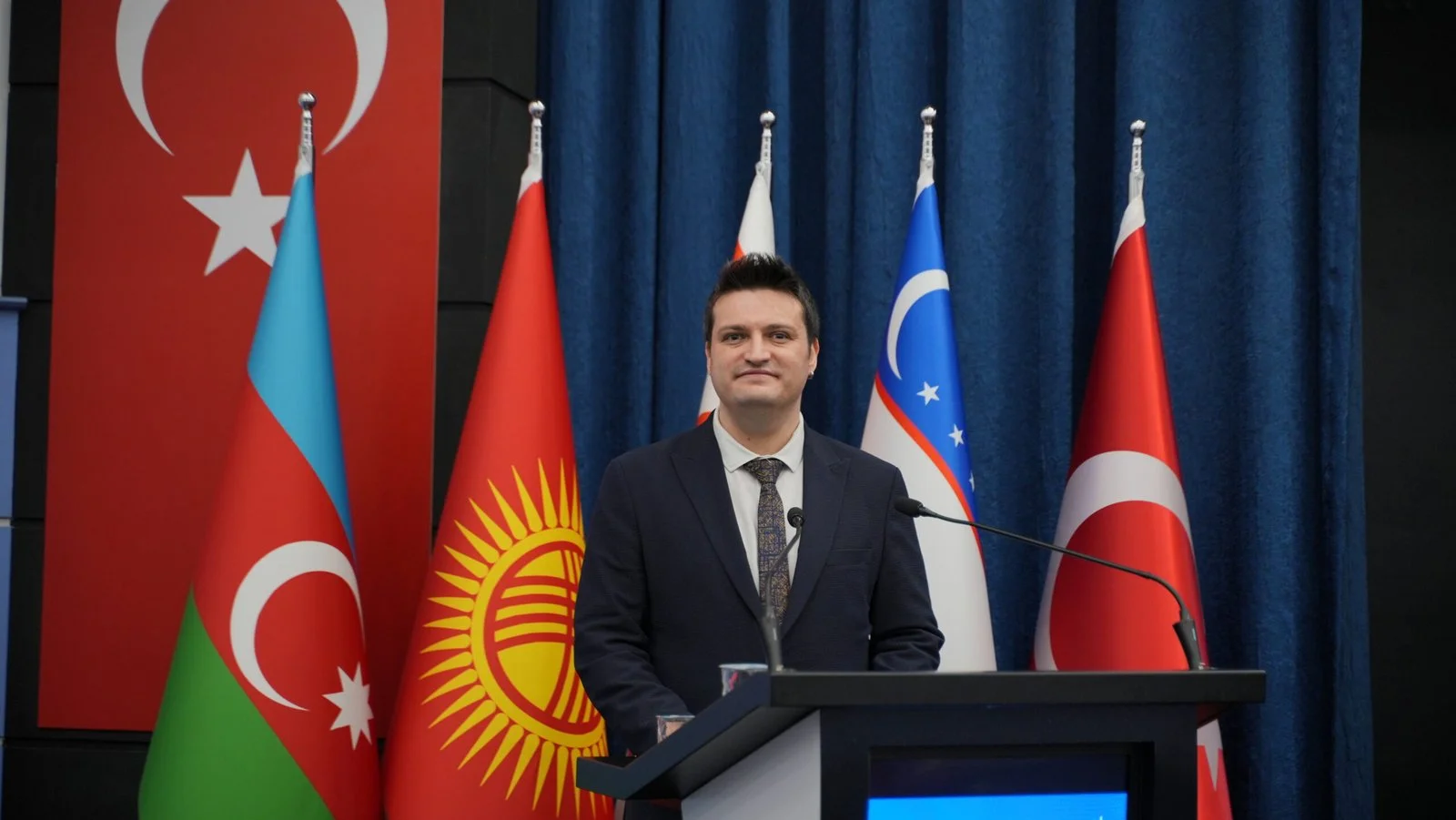 Türkiye, Azerbaycan, Kırgızistan ve Özbekistan bayrakları önünde kürsüde konuşan takım elbiseli genç bir adam