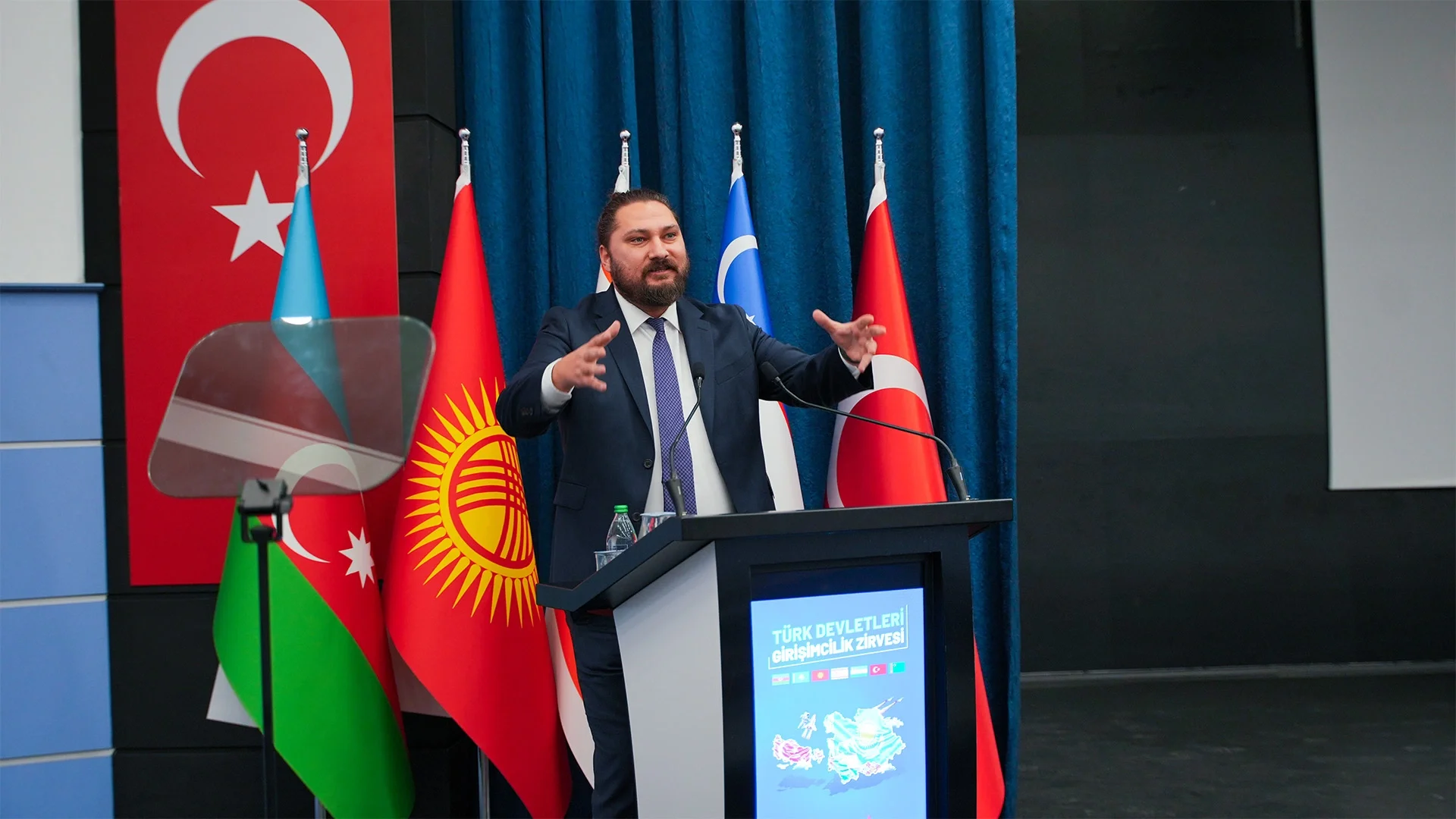 Türkiye, Azerbaycan, Kırgızistan ve Özbekistan bayrakları önünde zirve kürsüsünde ellerini açarak konuşan sakallı bir adam