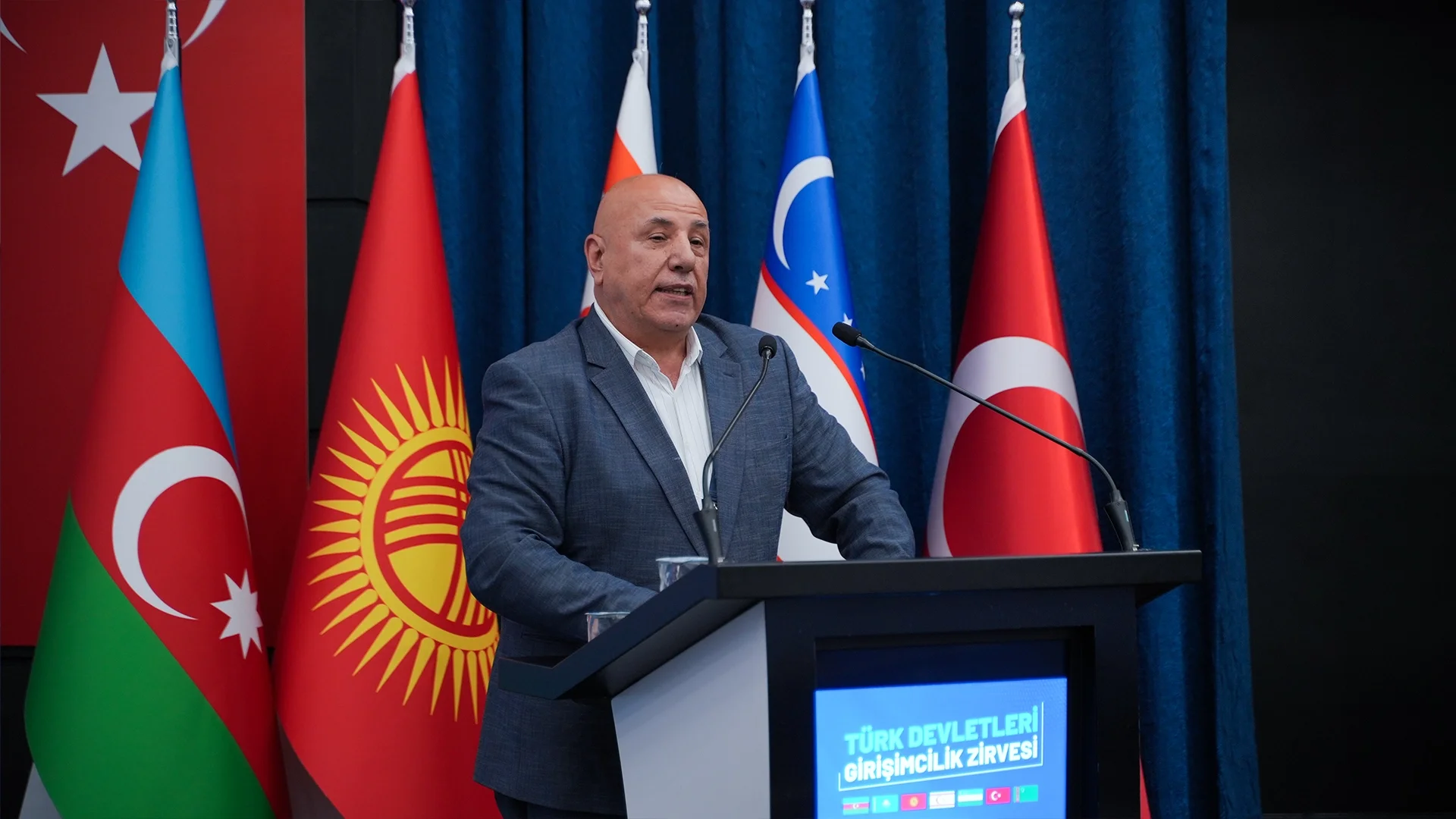 Azerbaycan, Kırgızistan, Özbekistan ve Türkiye bayrakları önünde, “Türk Devletleri Girişimcilik Zirvesi” yazılı kürsüde konuşan ceketli bir adam