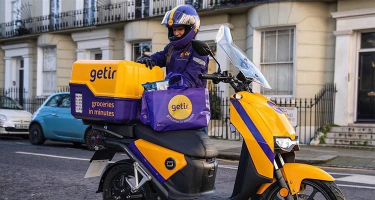 Tarihi binaların önündeki sokakta, arkasında getir kutusu bulunan sarı-mor elektrikli motosikletle teslimat yapan kask takmış kurye