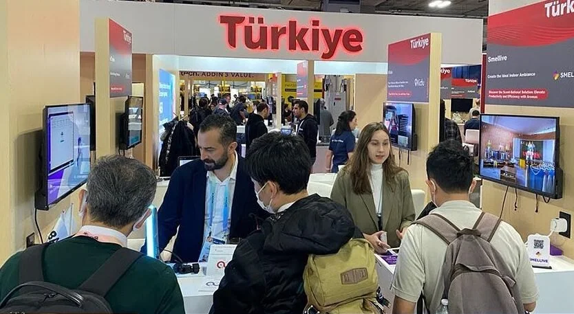 Fuarda “Türkiye” yazılı pavyonda ekranları ve ürünleri inceleyen ziyaretçiler ile stant görevlileri