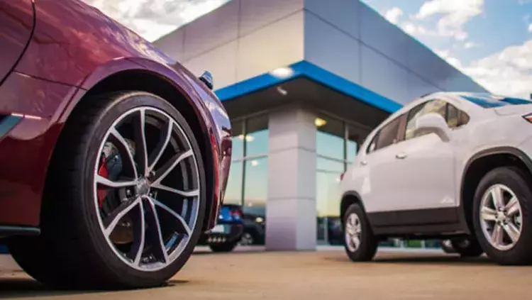 Bayi önünde park halindeki bordo bir otomobilin jantı ve arkada beyaz bir SUV