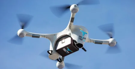 Mavi gökyüzünde uçan, altında logolu bir kutu taşıyan beyaz dört pervaneli drone