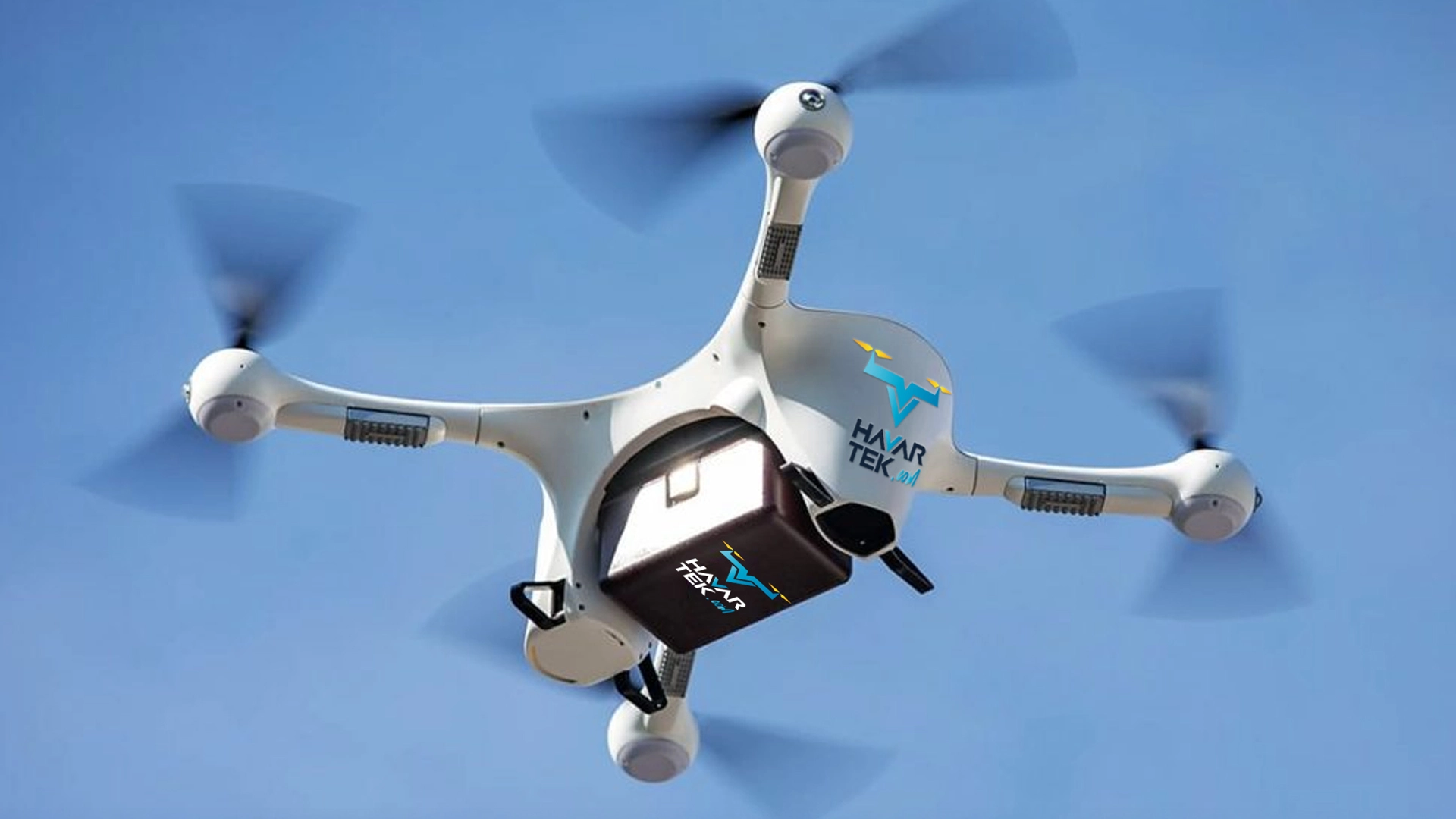 Mavi gökyüzünde uçan, altında logolu bir kutu taşıyan beyaz dört pervaneli drone