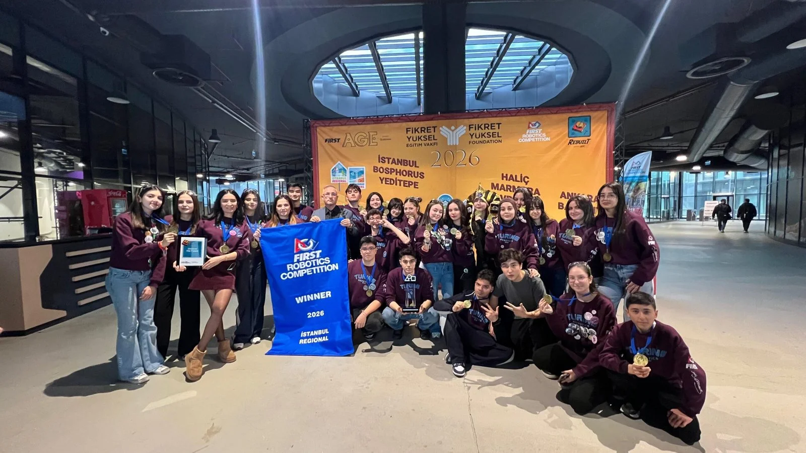 Bordo tişörtlü lise öğrencilerinden oluşan robotik takımı, FIRST Robotics Competition “Winner” bayrağı ve ödülüyle bir alışveriş merkezinde poz veriyor