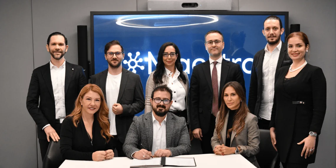 MaestroHub logolu ekran önünde, masada belge imzalayan bir adamın çevresinde poz veren ekip