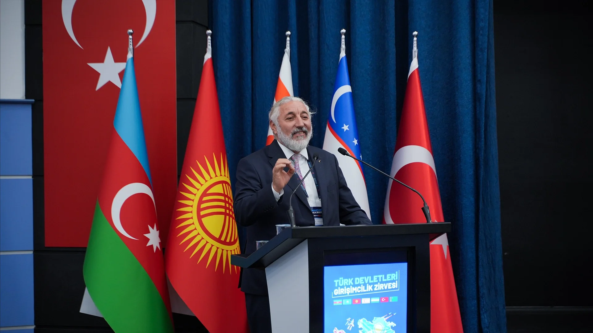 Türkiye, Azerbaycan, Kırgızistan ve Özbekistan bayrakları önünde, “Türk Devletleri Girişimcilik Zirvesi” kürsüsünde konuşan gözlüklü, sakallı bir adam