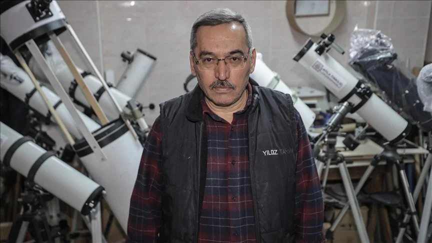Gözlüklü, ekose gömlekli ve yelekli bir adam, atölyede sıralanmış beyaz teleskopların önünde duruyor